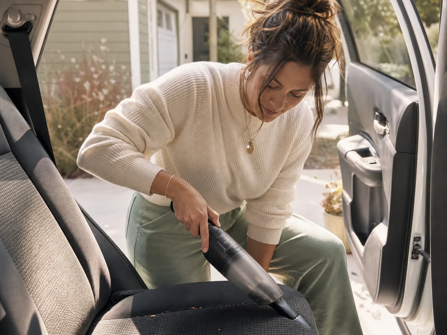 Woman using a compact handheld vacuum to clean crumbs from a car floor mat and seat crevice