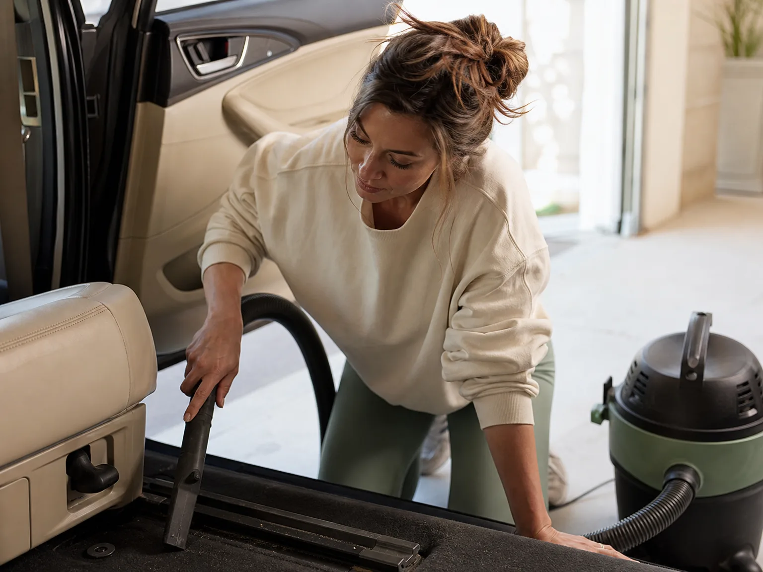 Woman using a shop vac hose to clean car seat rails in a sunlit garage