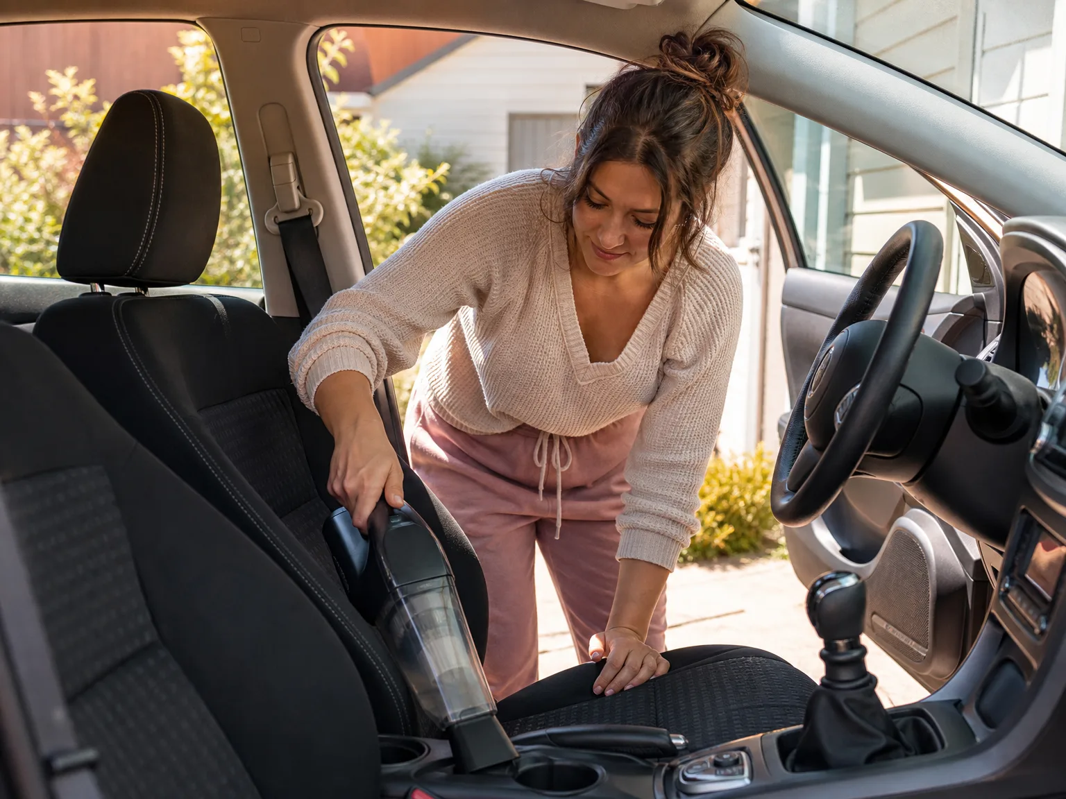 A woman in her early thirties using a cordless handheld vacuum on car seat crumbs in a sunlit driveway