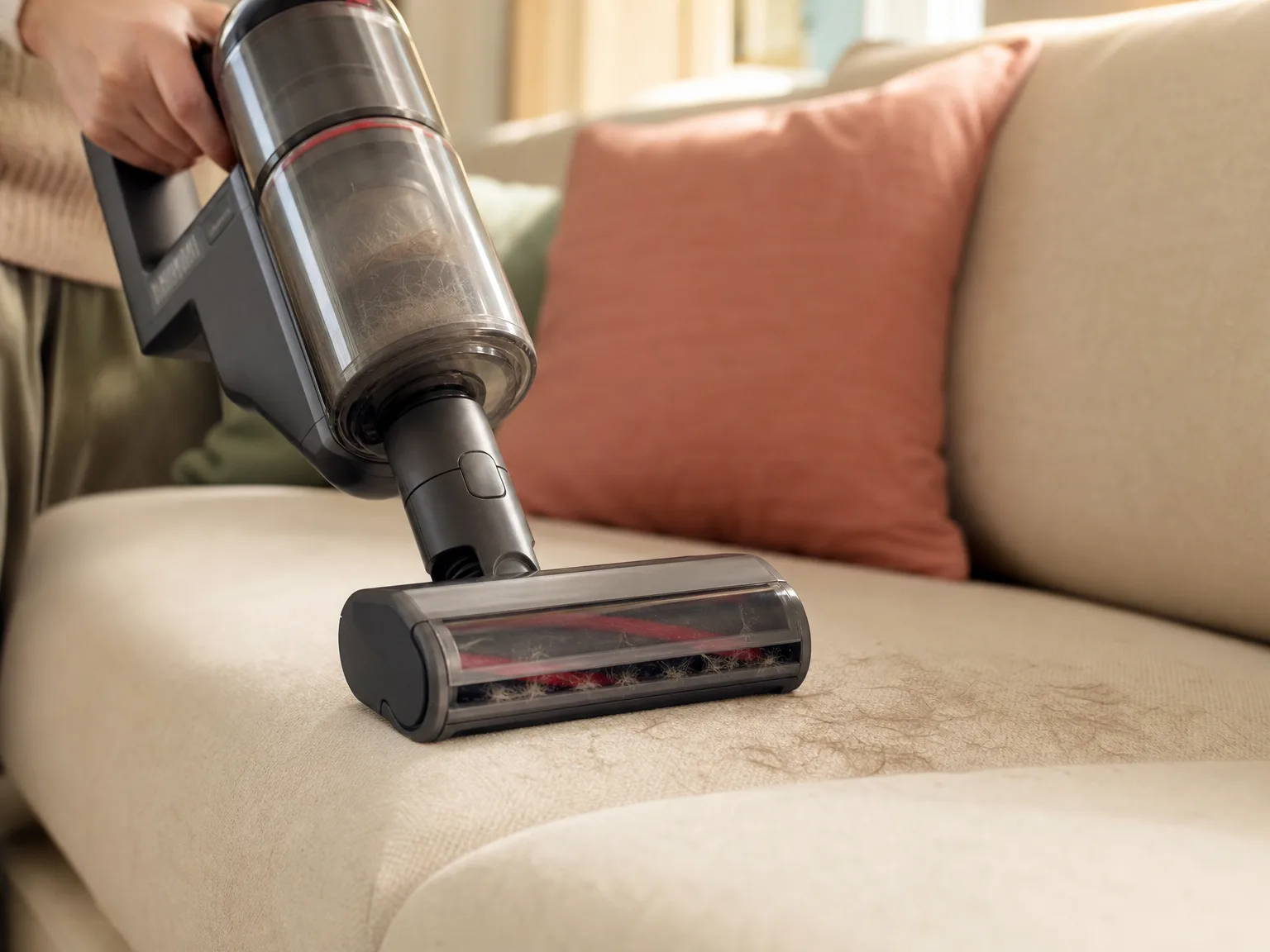 Close-up of a cordless handheld vacuum motorized rubber brush lifting cat fur from a cream sofa cushion in warm window light