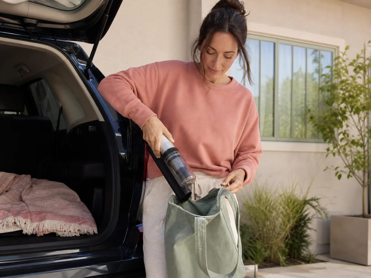 A woman in her early thirties packing a cordless handheld vacuum into a tote bag beside a car trunk in a sunlit driveway