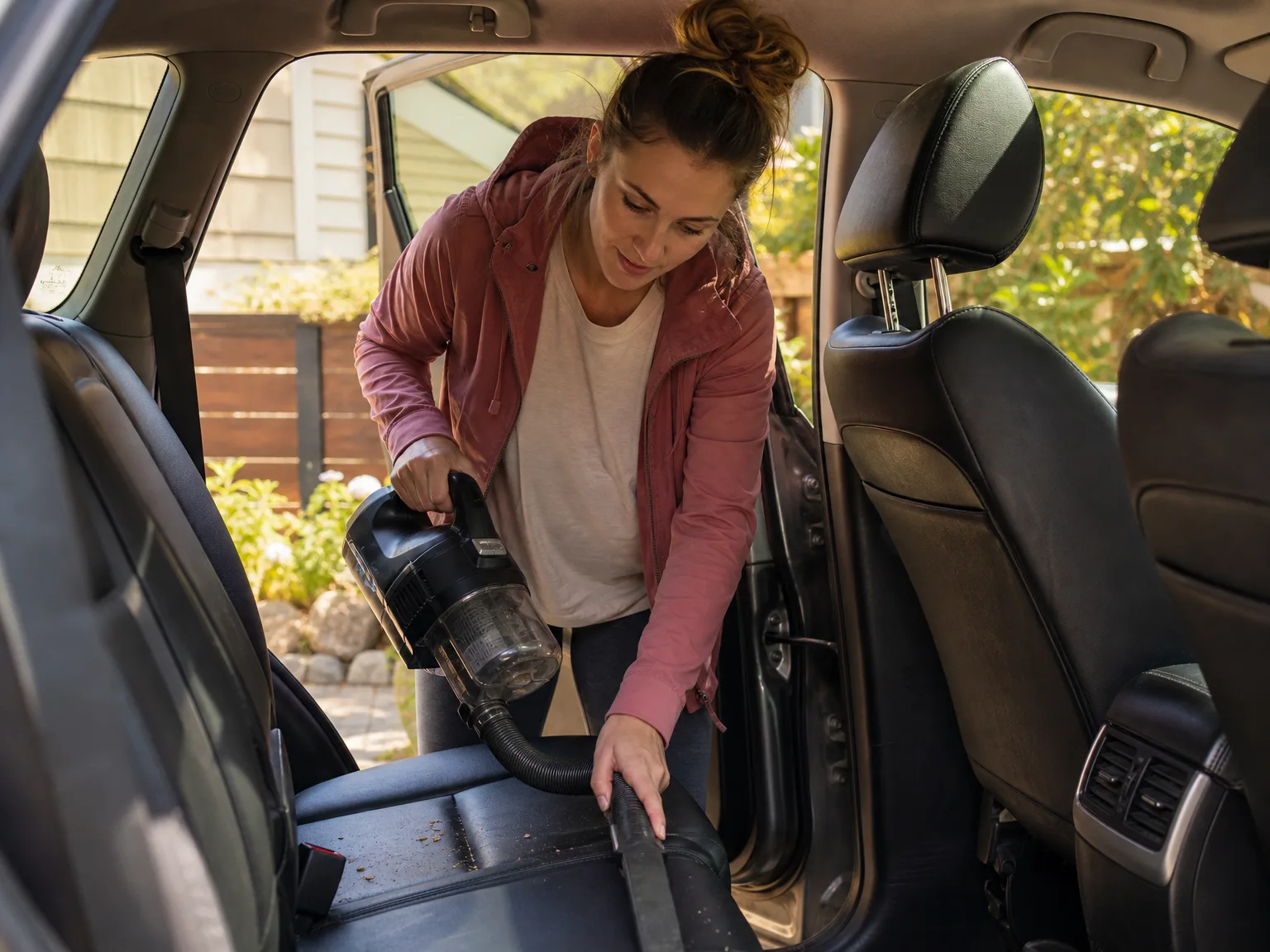 A woman in her early thirties using the EGO cordless shop vac to clean a car interior in her driveway, warm afternoon light
