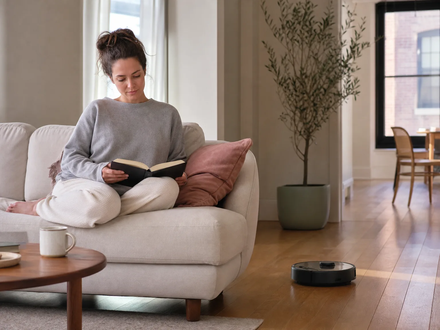 Woman reading on a cream sofa while a slim robot vacuum quietly cleans hardwood in a bright apartment
