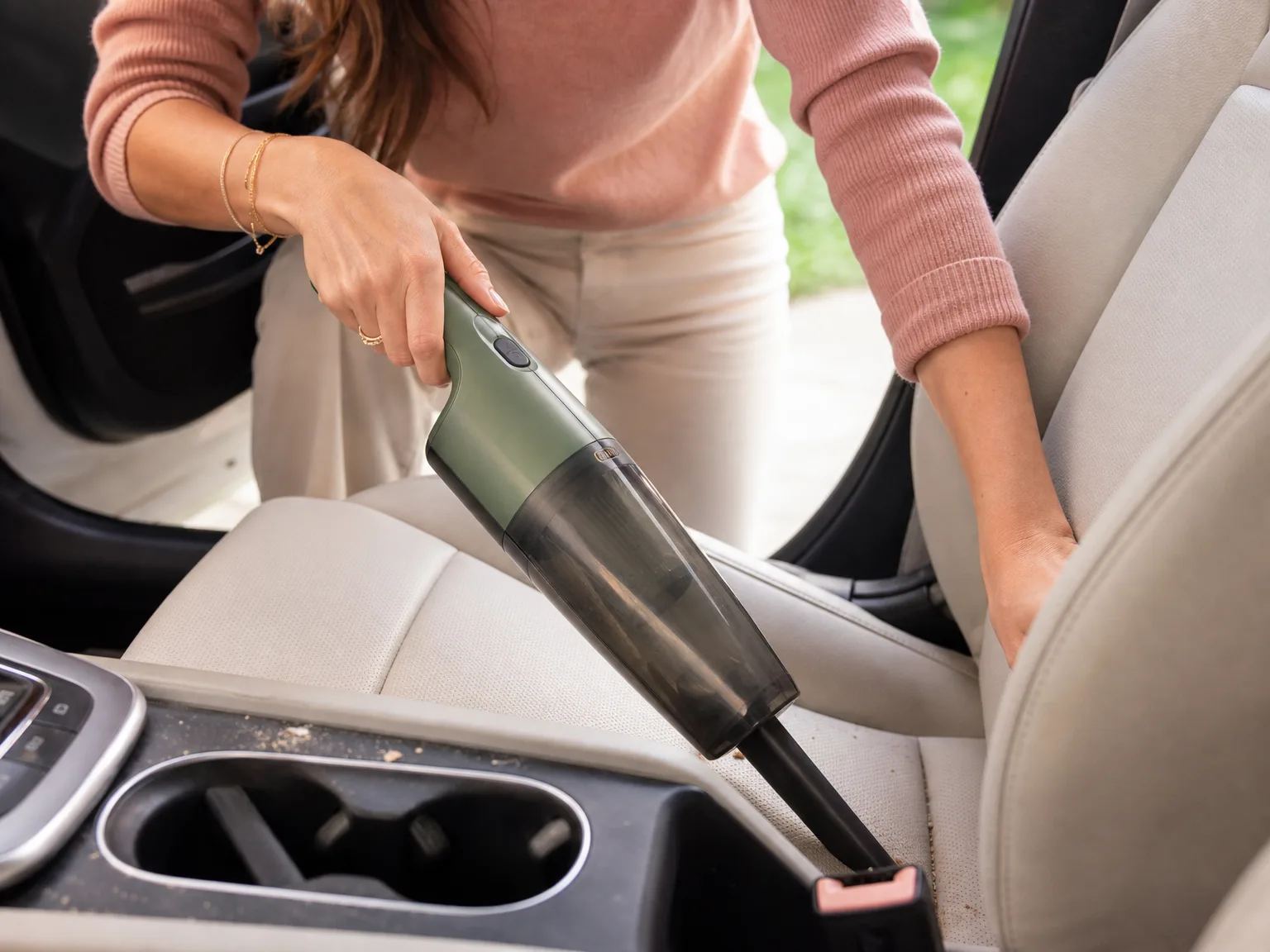 Hands guiding a slim handheld vacuum nozzle into a car seat crevice beside a dusty cup holder
