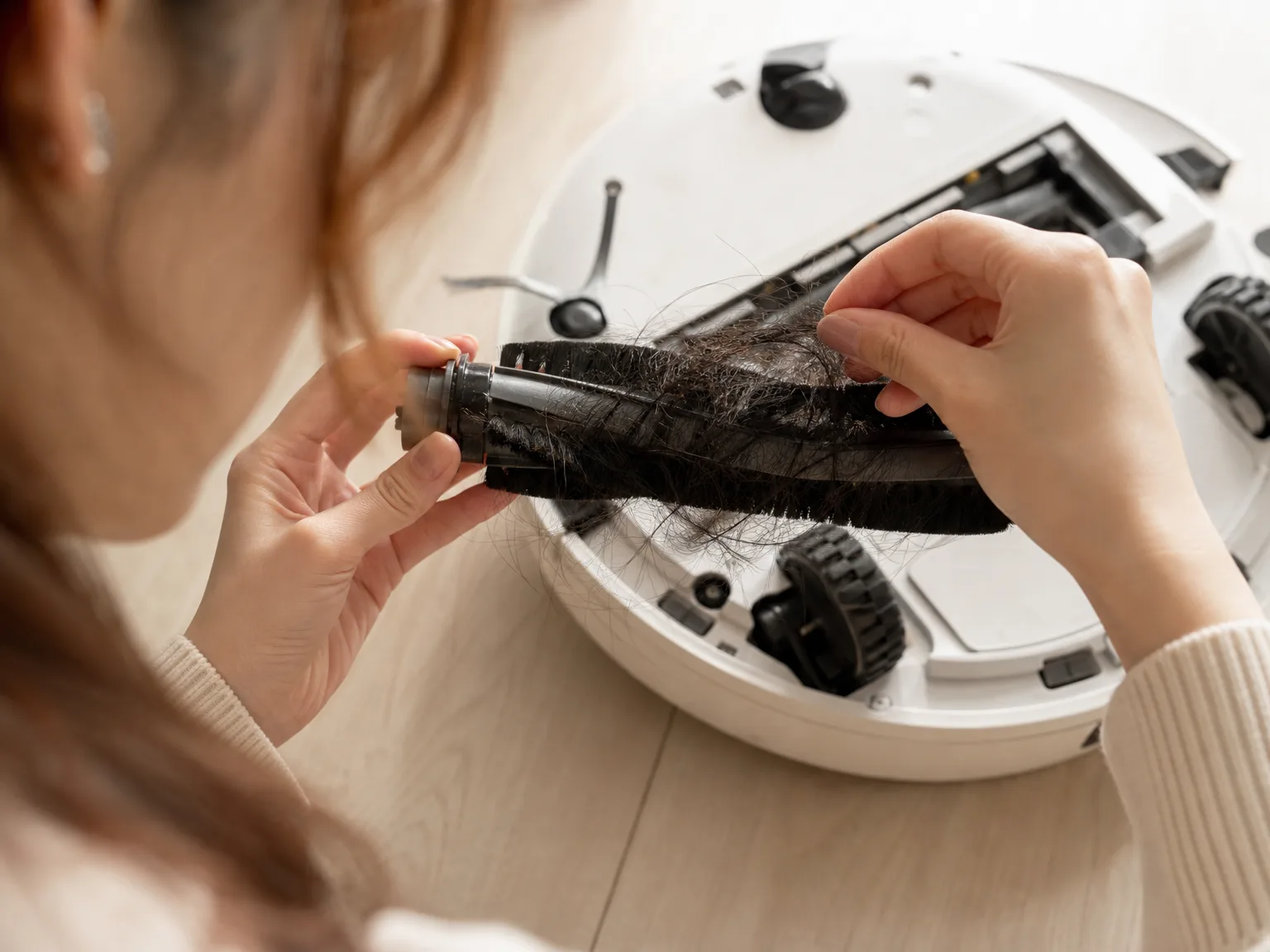 Close-up of a robot vacuum brush roller being cleared of long hair over a cream hardwood floor