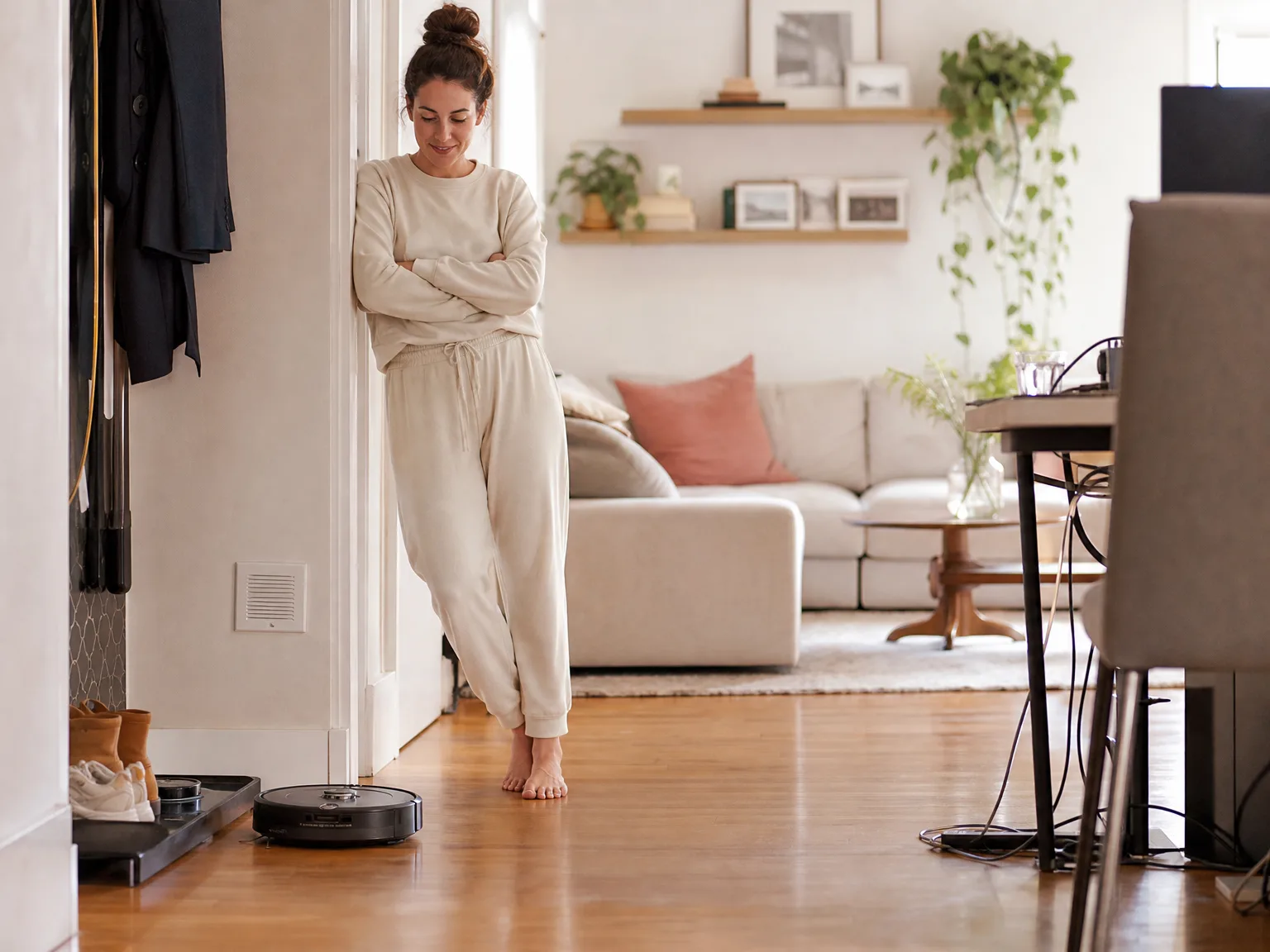 A woman in casual loungewear watching a round robot vacuum clean hardwood near a shoe rack and charging cables in a sunlit living room
