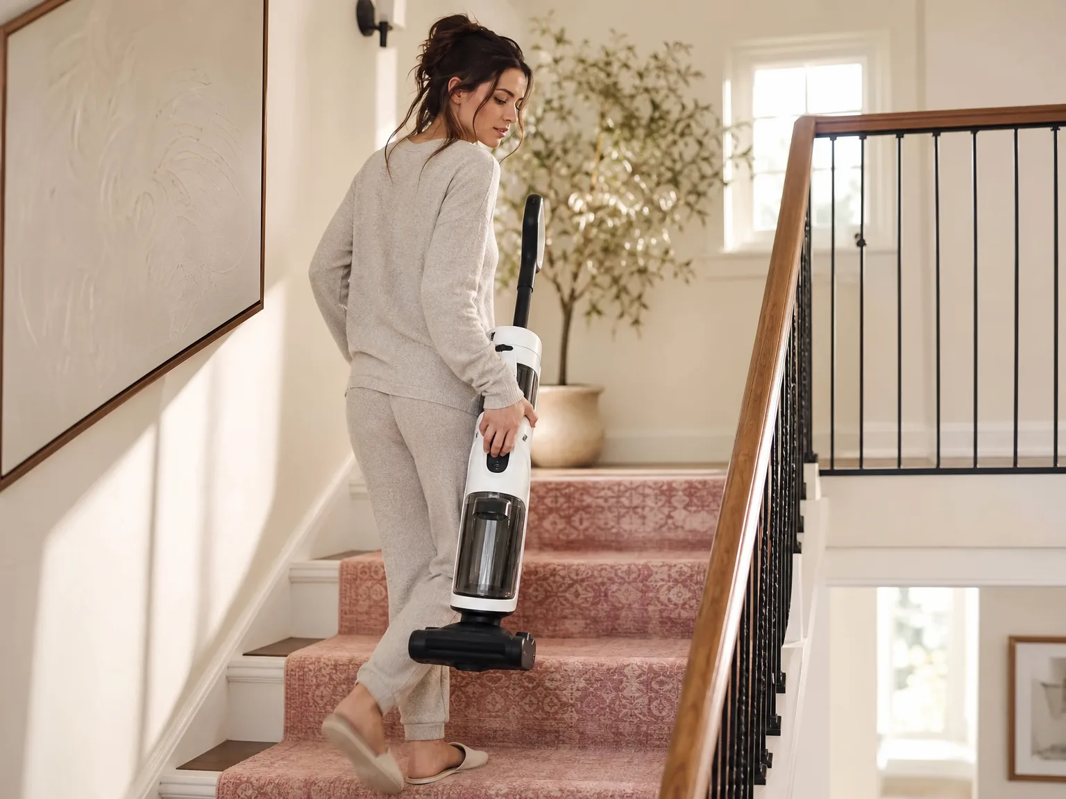 Woman carrying a slim lightweight upright vacuum up a sunlit hallway staircase in a lived-in home