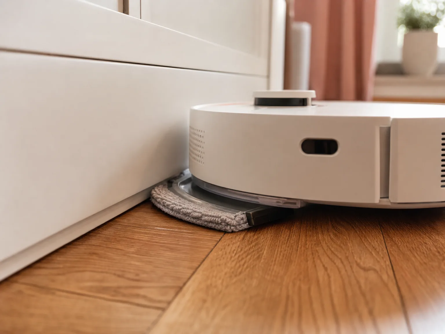 Close-up of a robot mop pad reaching along a baseboard and cabinet toe kick on warm hardwood in window light