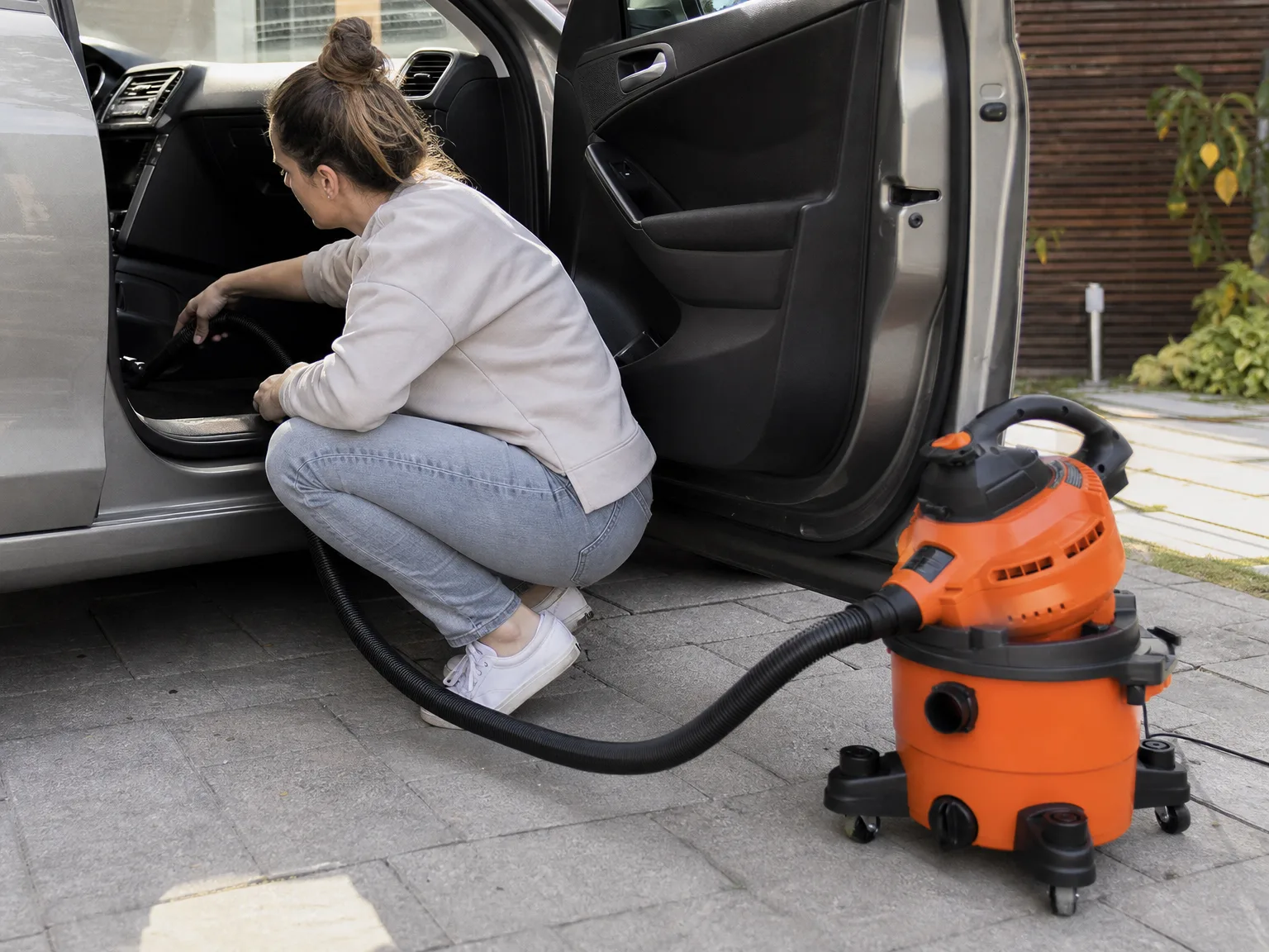 Woman in her thirties crouching beside an open car door in a driveway, vacuuming sand out of the footwell with a wet dry vacuum hose