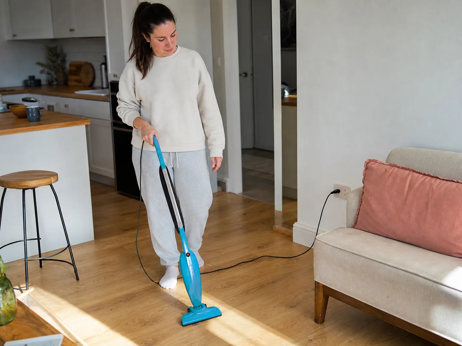 Woman in her early thirties running a slim blue stick vacuum across sunlit hardwood in a small studio apartment kitchen and living area