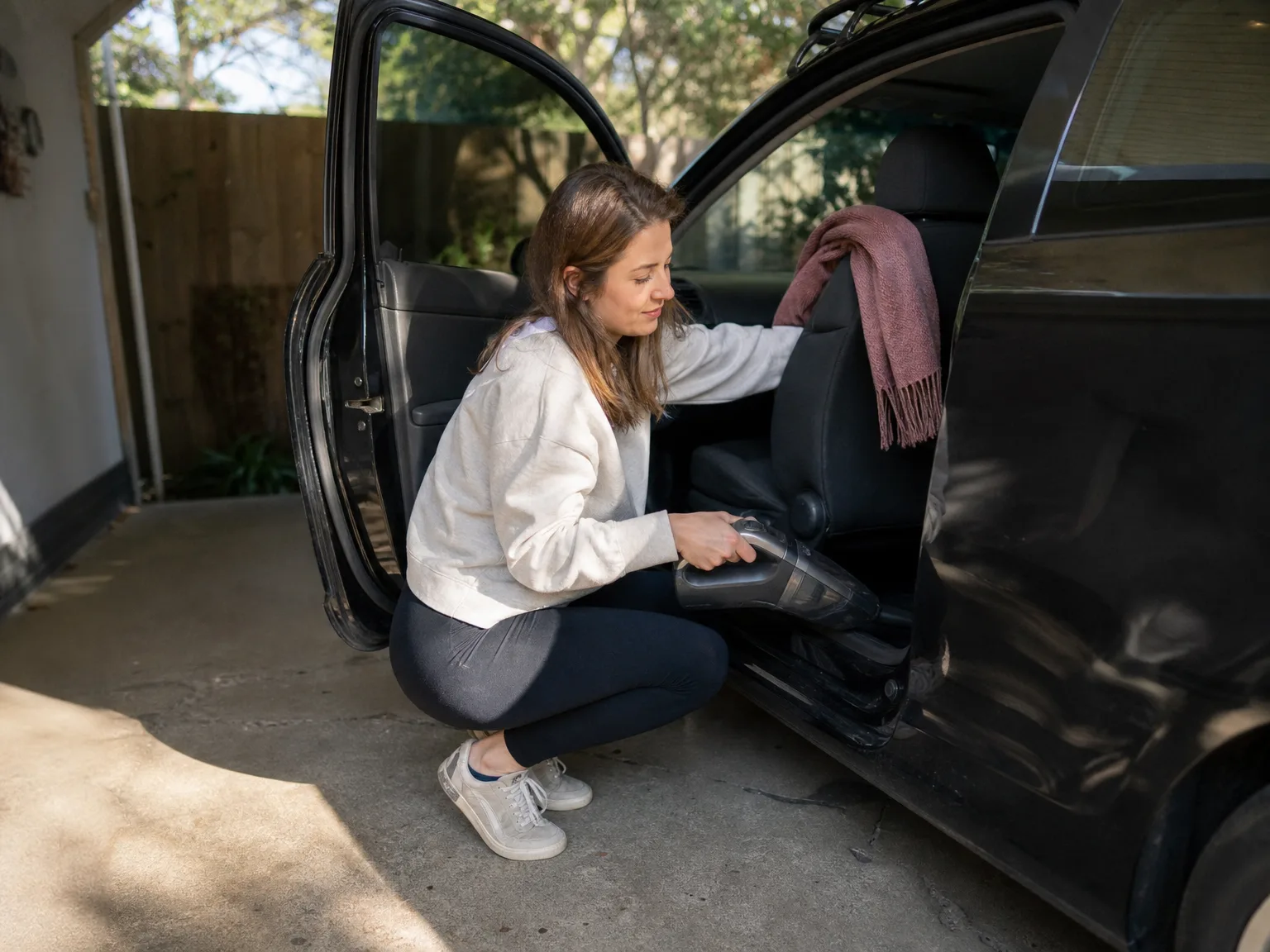 Woman in her early thirties crouching beside an open car door in a sunlit driveway, cleaning the passenger seat with a compact cordless handheld vacuum