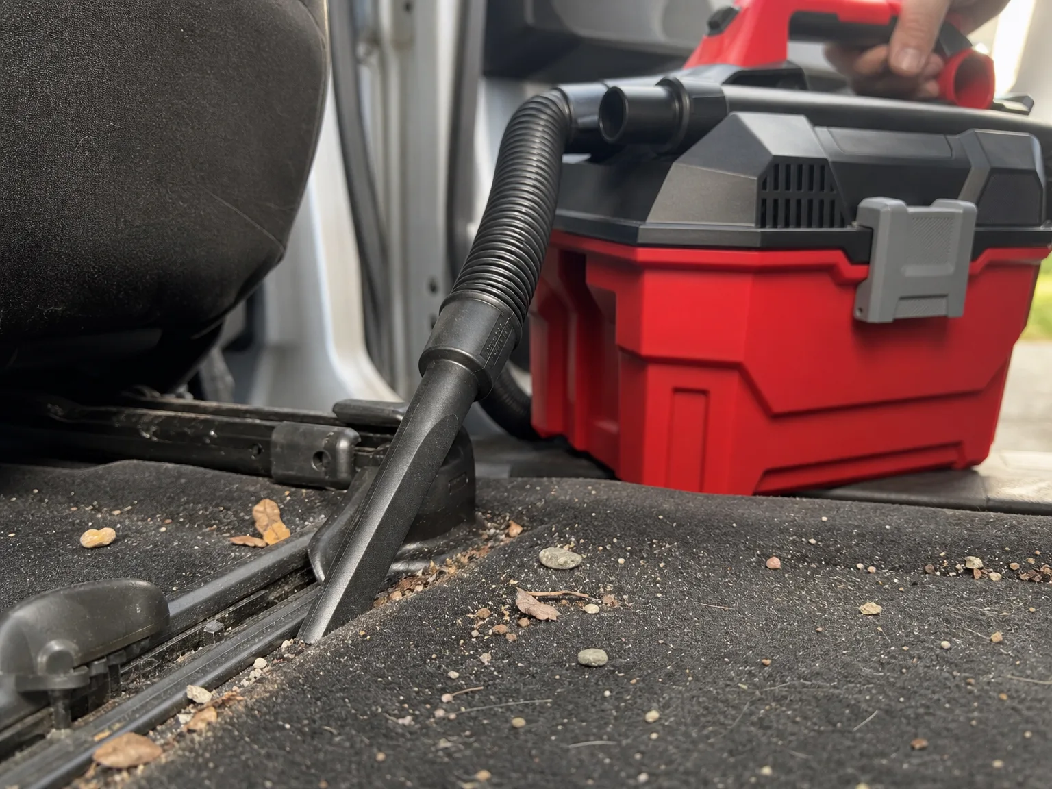 Close-up of a cordless wet/dry vac crevice nozzle pulling sand and grit out of the seat rail track in a car footwell