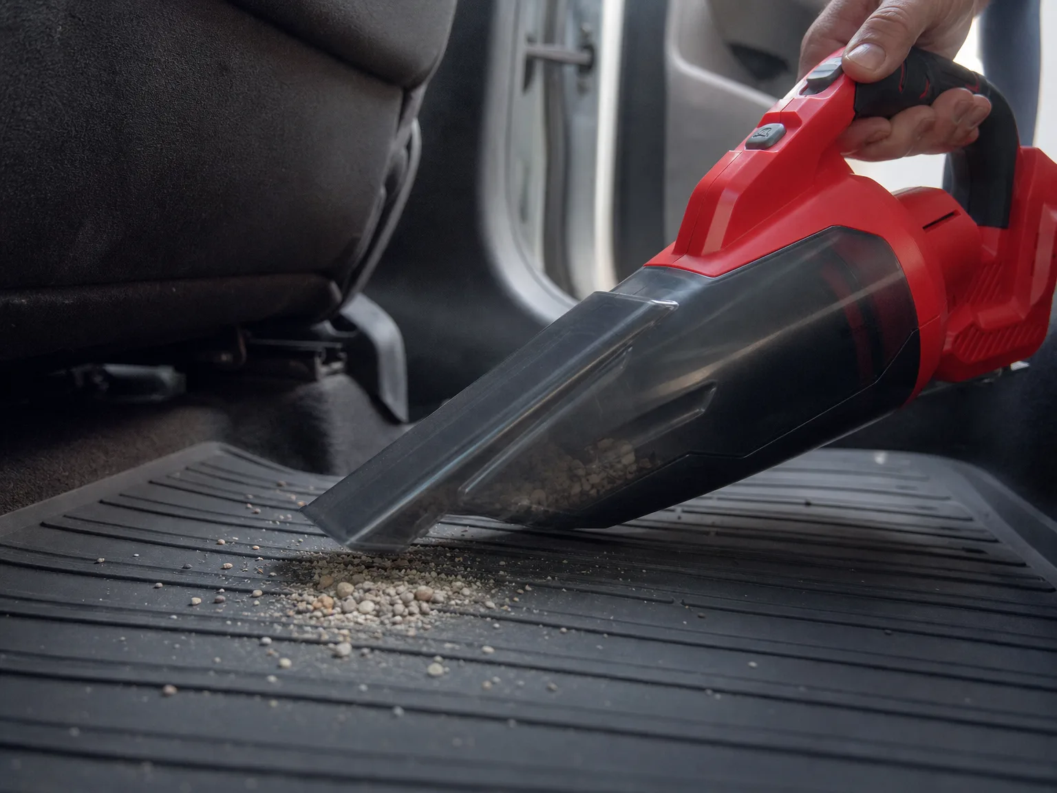 Close-up of a cordless hand vacuum nozzle lifting fine gravel and grit from the ribbed rubber floor mat of a car footwell