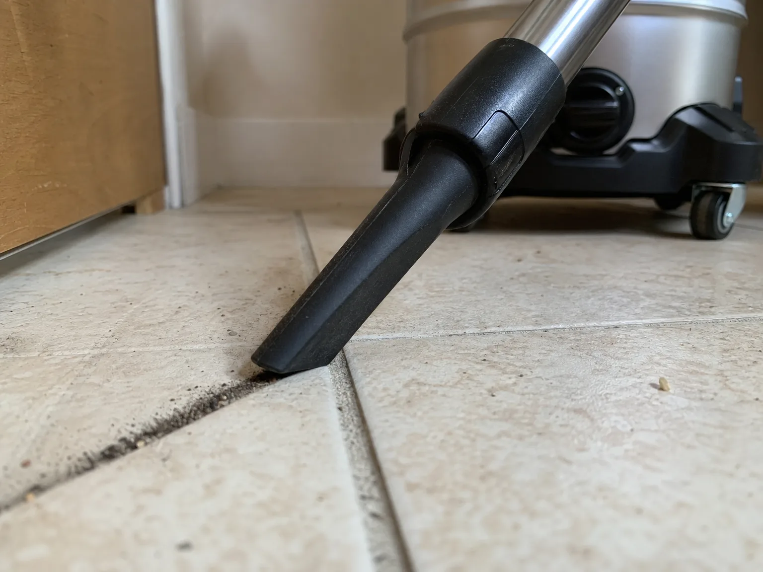 Close-up of a shop vacuum crevice nozzle pulling dark grit out of the grout line between pale floor tiles