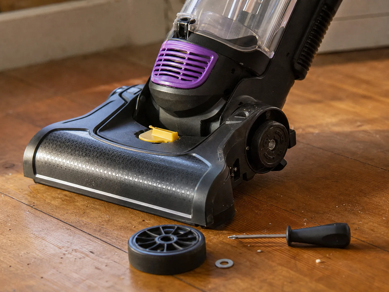 Rear wheel of an upright vacuum sitting loose on a hardwood floor beside the tilted machine with a small screwdriver next to it