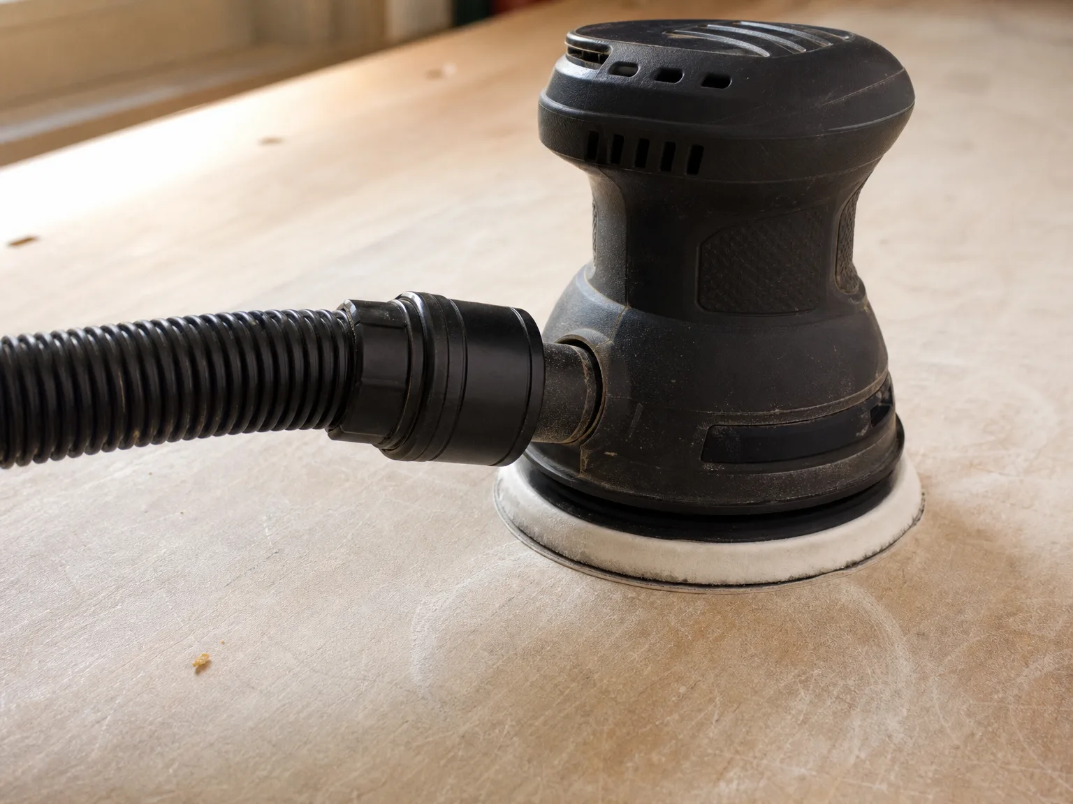 Close-up of a dust extractor hose cuff clamped onto the dust port of a random orbit sander on a plywood workbench