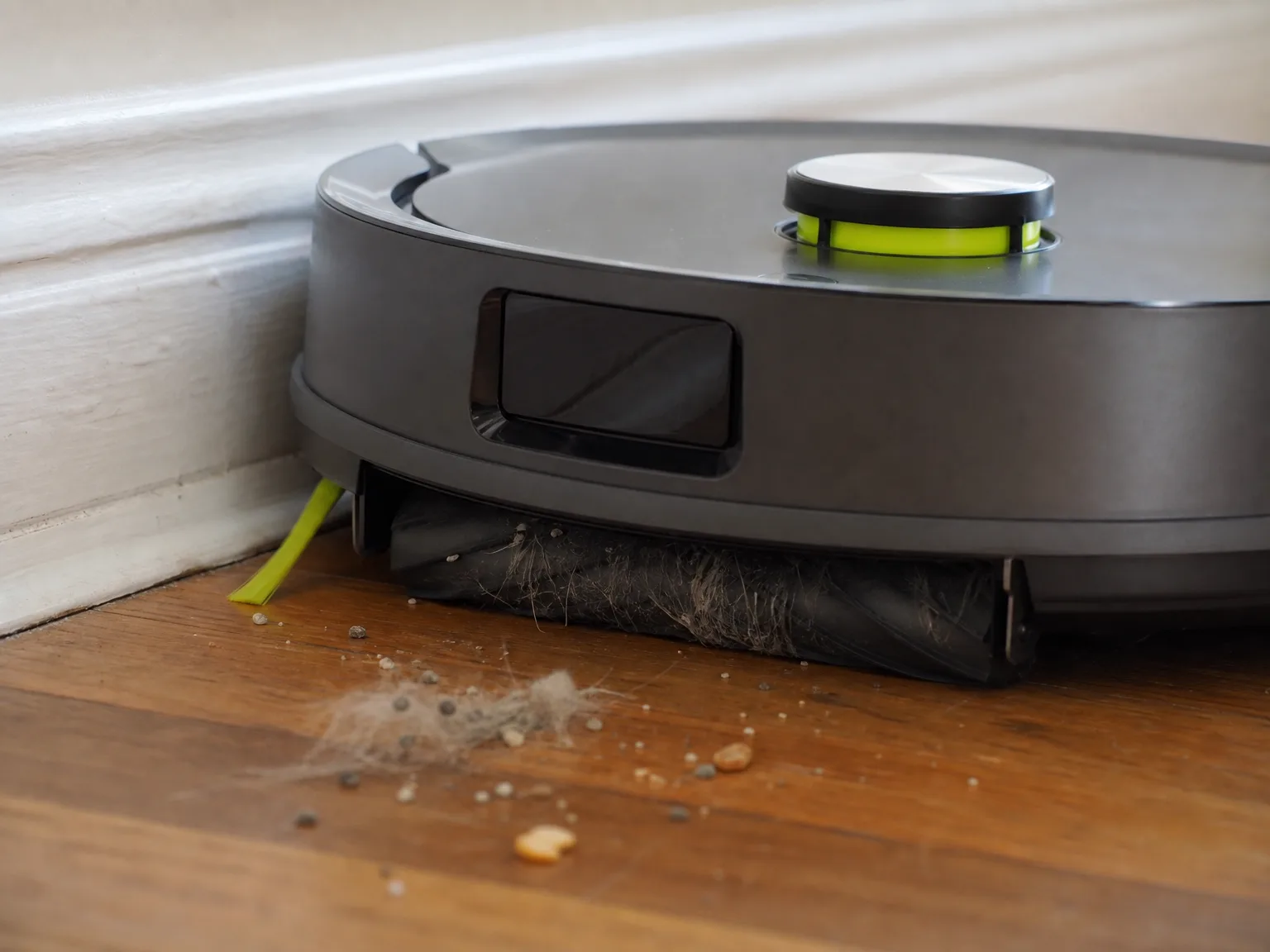 Close-up of a robot vacuum's anti-tangle roller lifting grey cat hair and litter granules from warm hardwood flooring beside a painted baseboard