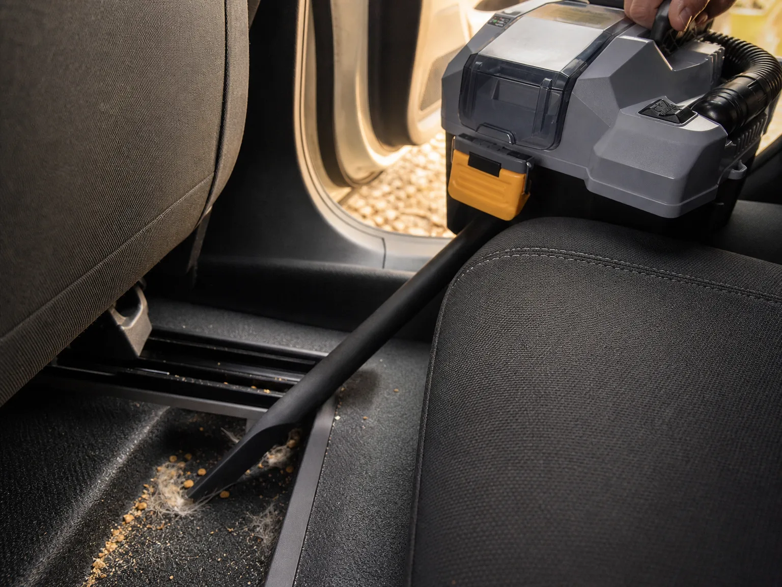 Close-up of a cordless shop vac crevice tool lifting sand and pet hair from the seat rail of a car interior