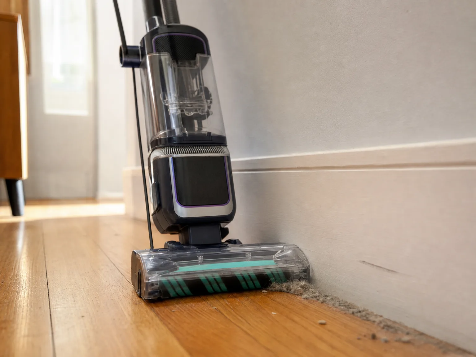 Close-up of an upright vacuum floor head pressed flush against a white baseboard, lifting a line of gray dust and pet hair from oak flooring