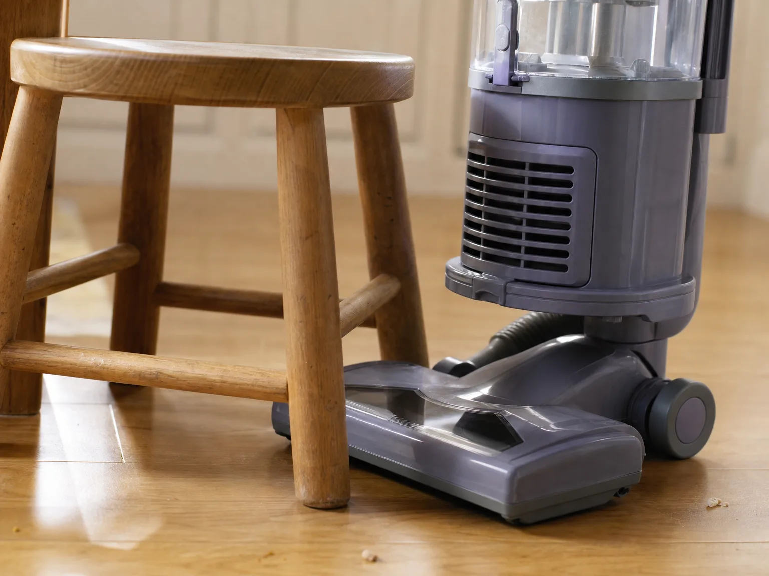 A wide upright vacuum floor head stopped at the edge of a low wooden chair, unable to fit into the gap beneath the seat