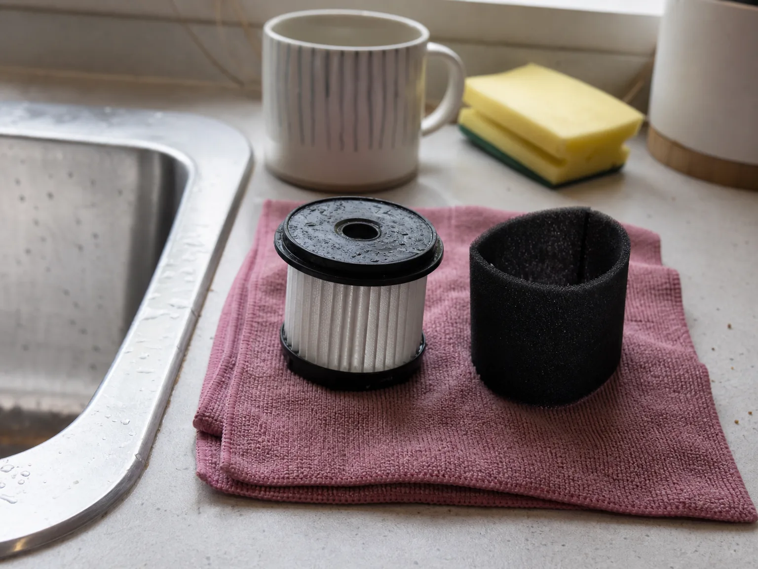 Two rinsed vacuum filters, a pleated round filter and a black foam sleeve, air-drying on a folded dusty rose towel beside a kitchen sink
