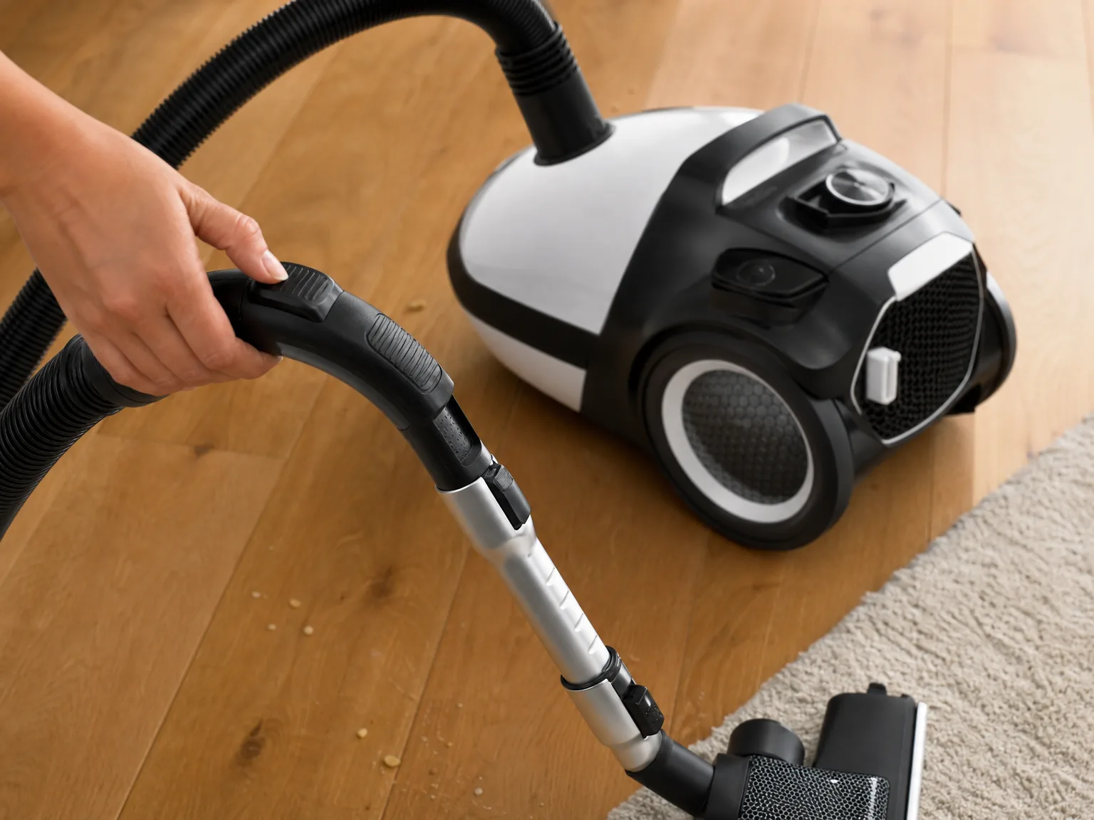 Close-up of a hand adjusting the suction slider on a canister vacuum wand handle above oak flooring, warm side window light