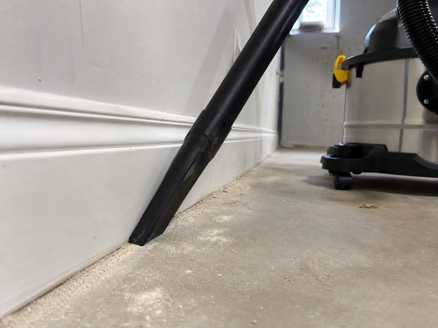 Close-up of a shop vacuum crevice tool pulling fine drywall dust out of the gap between a basement floor and the baseboard