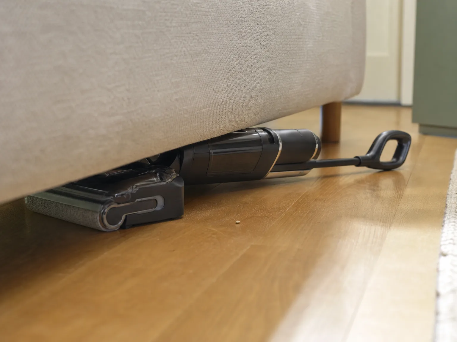A wet dry vacuum cleaner reaching flat under a low sofa to clean hidden dust on a warm hardwood floor in natural window light