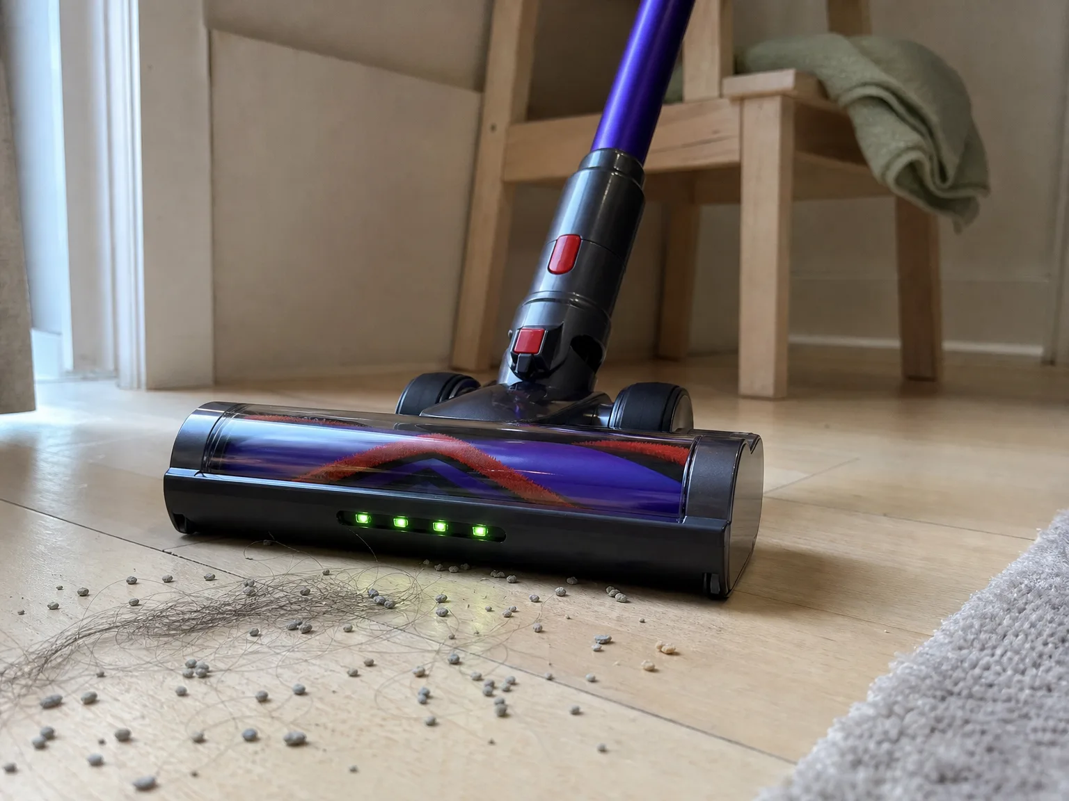 Close-up of a cordless vacuum floor head lifting strands of pet hair and scattered cat litter from warm cream hardwood next to a low-pile rug